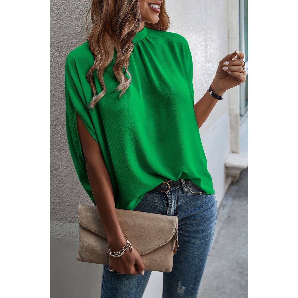 Woman wearing a green blouse and blue jeans, holding a beige clutch against a gray wall.