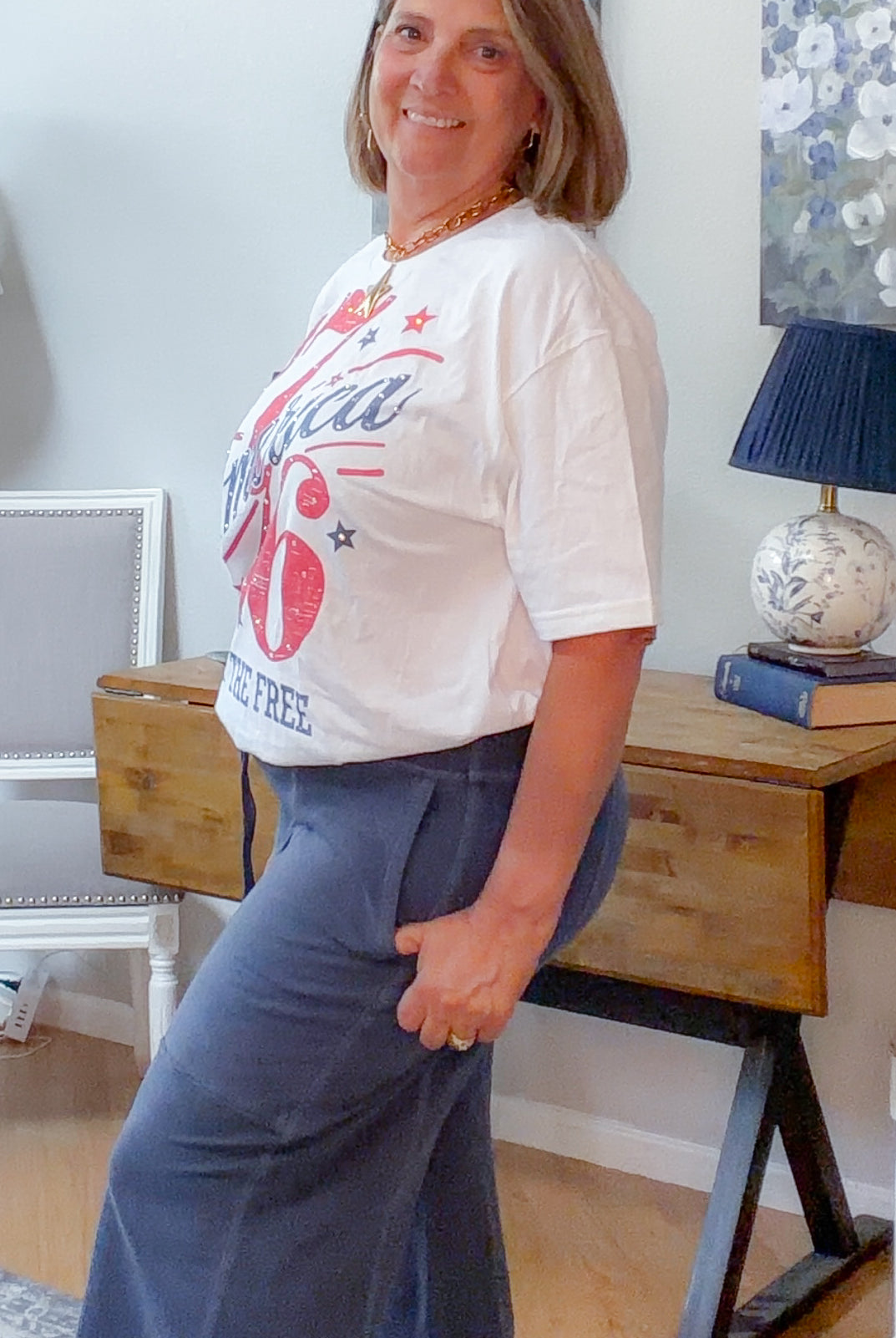 Woman wearing a white t-shirt with red graphics and blue pants in a room with a desk and lamp.