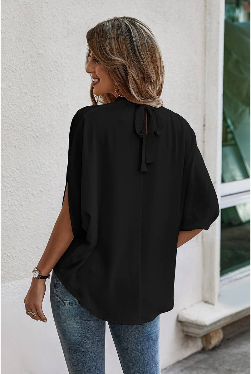 Woman wearing a black blouse with a bow detail and blue jeans, standing against a white wall.