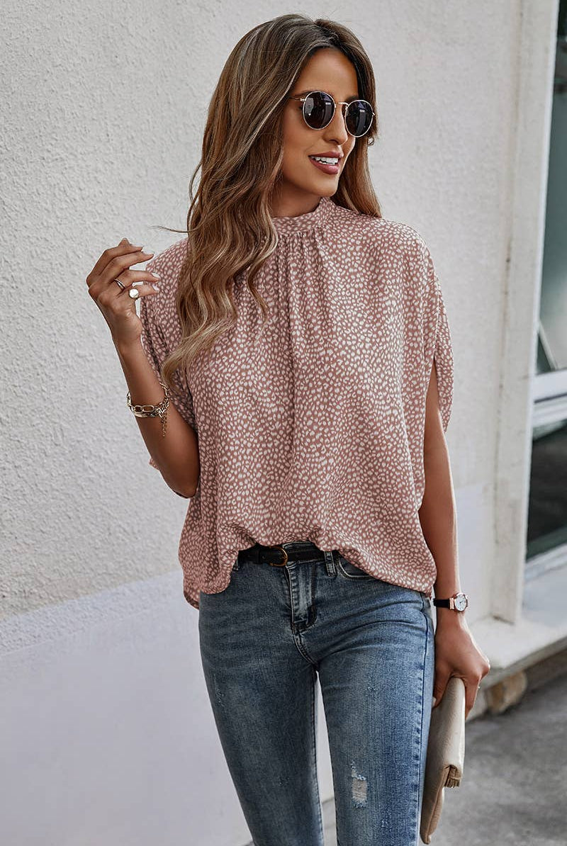 Woman wearing a patterned blouse and jeans against a white wall.