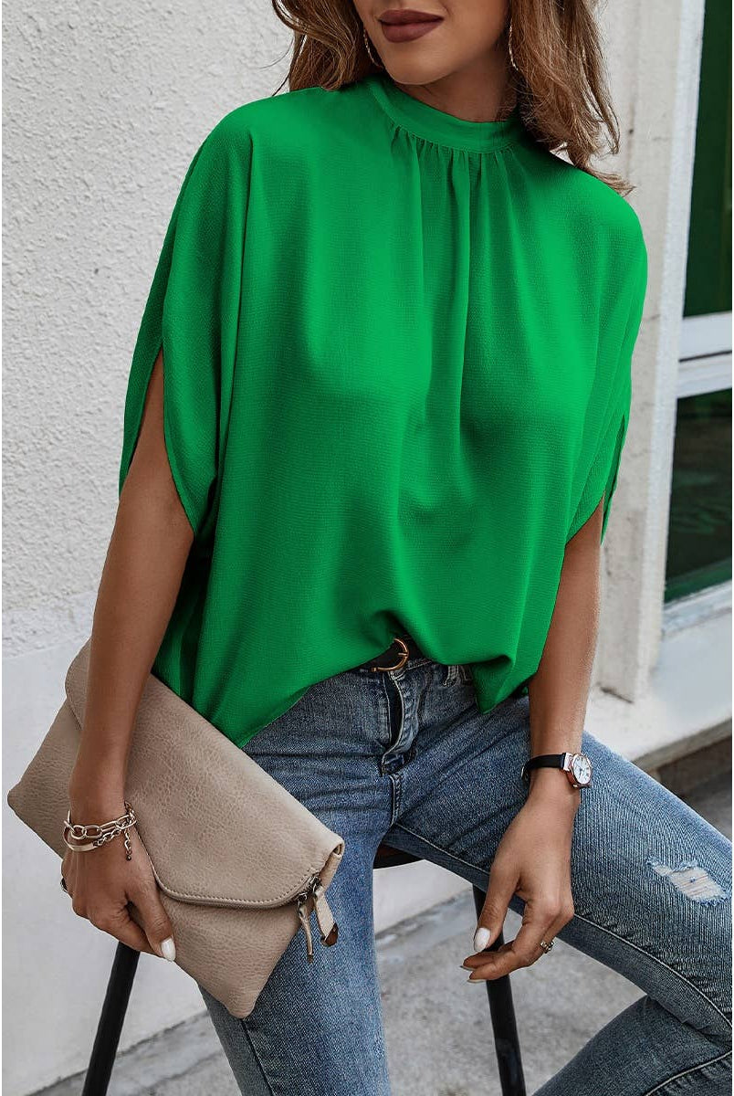 Person wearing a green blouse and blue jeans, holding a beige clutch against a white wall.