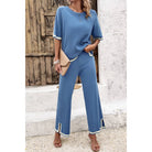 Woman wearing a blue outfit with white trim standing against a rustic wooden door and white wall.