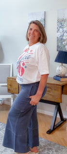 Woman wearing a white t-shirt with red graphics and blue pants in a room with a desk and lamp.