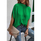 Person wearing a green blouse and blue jeans, holding a beige clutch against a white wall.