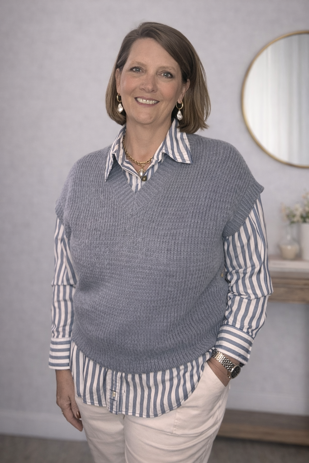 Woman wearing a gray sweater vest over a striped shirt in a room with a mirror on the wall.